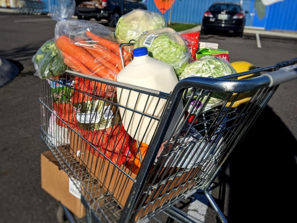 A shopping cart full of groceries including bagged and boxed items, fresh produce, and milk, stands in a parking lot at OneGenAway's mobile food pantry.