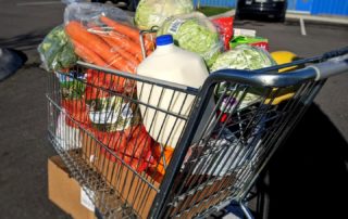 A shopping cart full of groceries including bagged and boxed items, fresh produce, and milk, stands in a parking lot at OneGenAway's mobile food pantry.