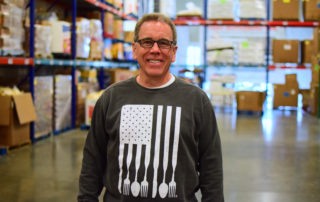 Chris Whitney, founder of OneGenAway, stands in OneGenAway's warehouse, which is full of pallets of cans and packaged food items.