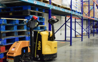 Two pallet jacks sit beside empty blue wooden pallets, and shelves along a wall in the OneGenAway warehouse sit empty.