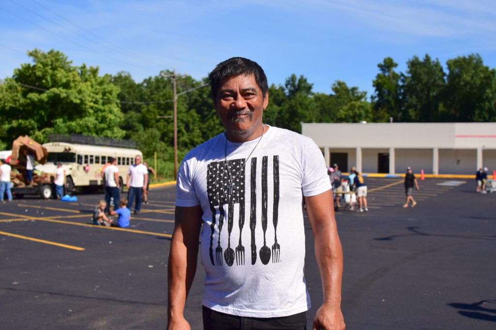 Steve, a man in a white OneGenAway T-shirt, smiles for the camera in front of the OneGenAway mobile food pantry in an open parking lot.