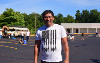 Steve, a man in a white OneGenAway T-shirt, smiles for the camera in front of the OneGenAway mobile food pantry in an open parking lot.