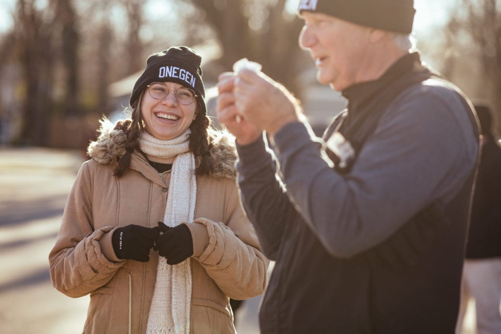 Tori Keafer, a woman in a puffy coat, black beanie, and braids, laughs as she talks with a man in a fleece vest and beanie.