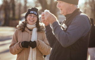 Tori Keafer, a woman in a puffy coat, black beanie, and braids, laughs as she talks with a man in a fleece vest and beanie.