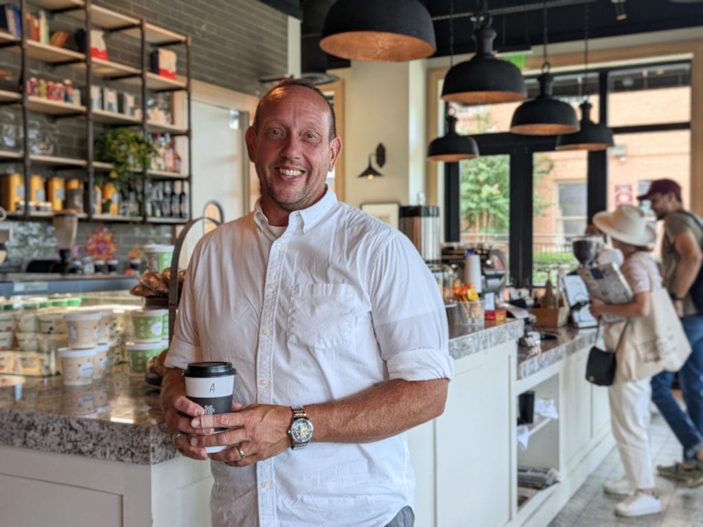 Richard Barnes, a man in a white short-sleeved dress shirt, smiles in front of a coffee bar