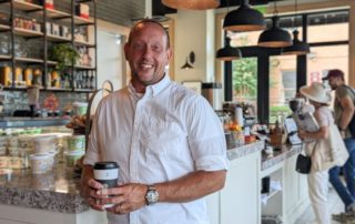 Richard Barnes, a man in a white short-sleeved dress shirt, smiles in front of a coffee bar